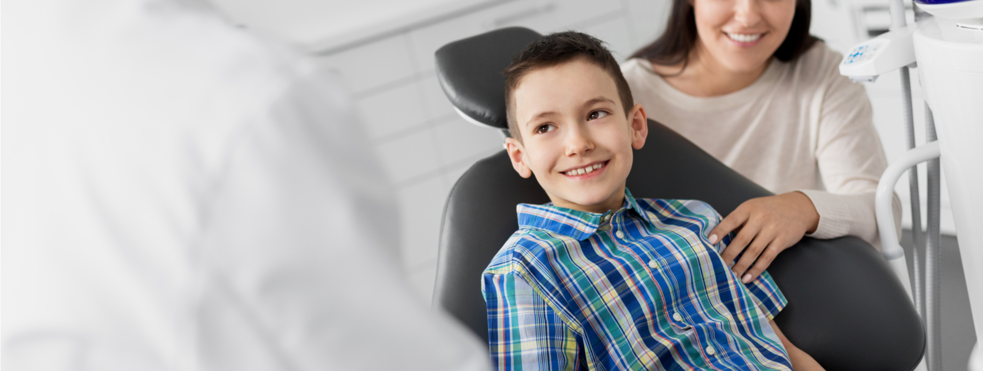 Kid Boy in Plaid Shirt Sitting in a Dental Office Chair With Mom Smiling at Dentist