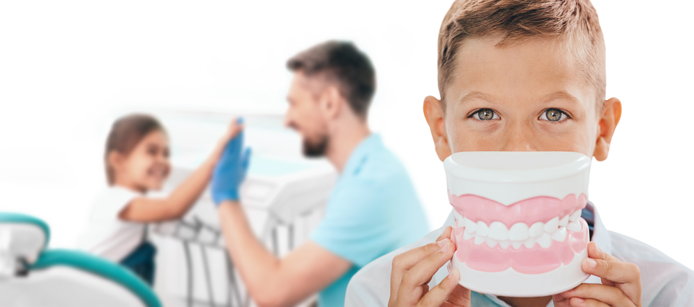 Kid Boy Holding Model Teeth Infront of His Smile With Sister and Dentist High-Fiving in the Backkground